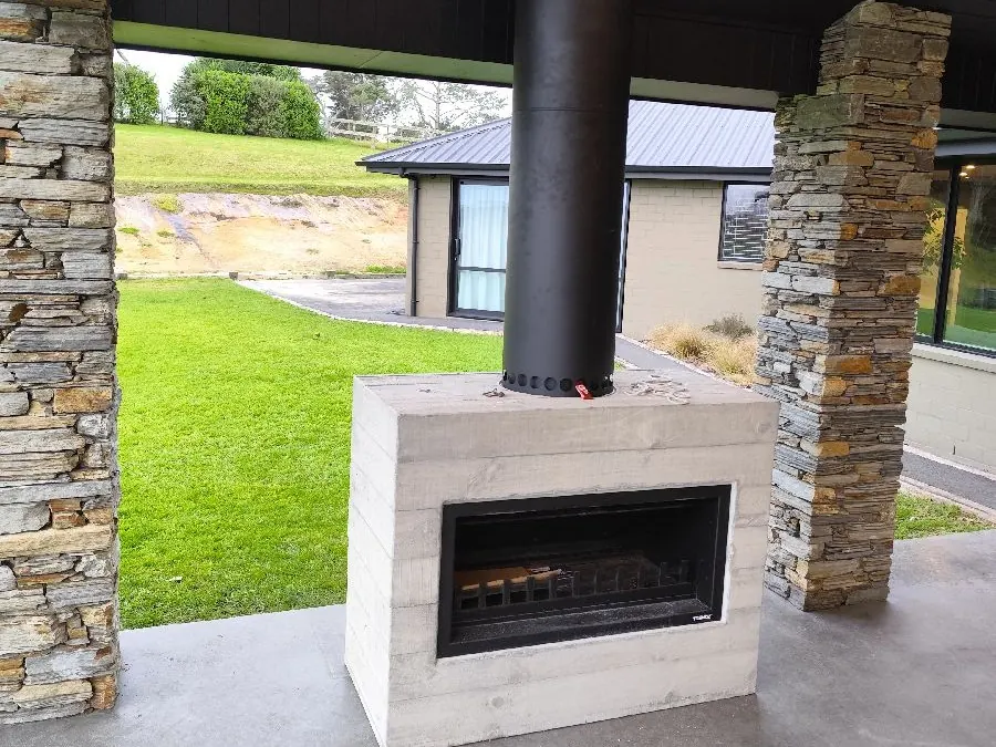 An outdoor wood fire in a concrete surround under a covered patio, with stone pillars and a black flue